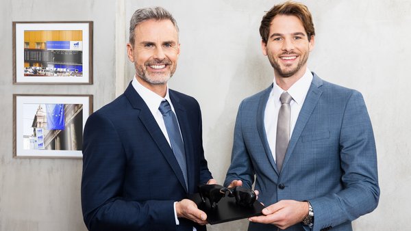 Dwóch mężczyzn w garniturach trzyma wspólnie rzeźbę przedstawiającą byka i niedźwiedzia – symbolizującą rynki finansowe i partnerską współpracę.