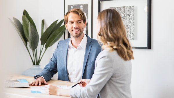 Mężczyzna w niebieskiej marynarce rozmawia przyjaźnie z kobietą w jasnym blezerze przy biurku.