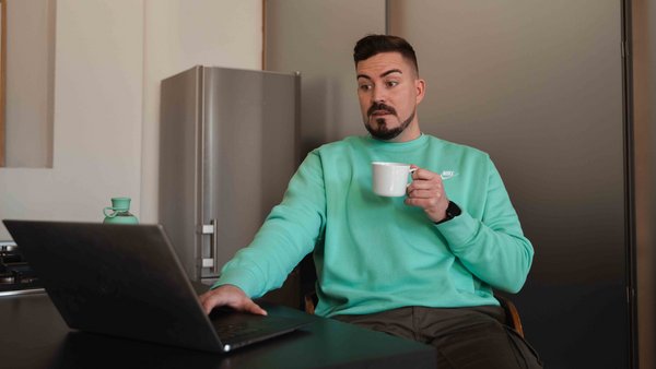 Mężczyzna siedzi zszokowany przed laptopem z kubkiem w dłoni.